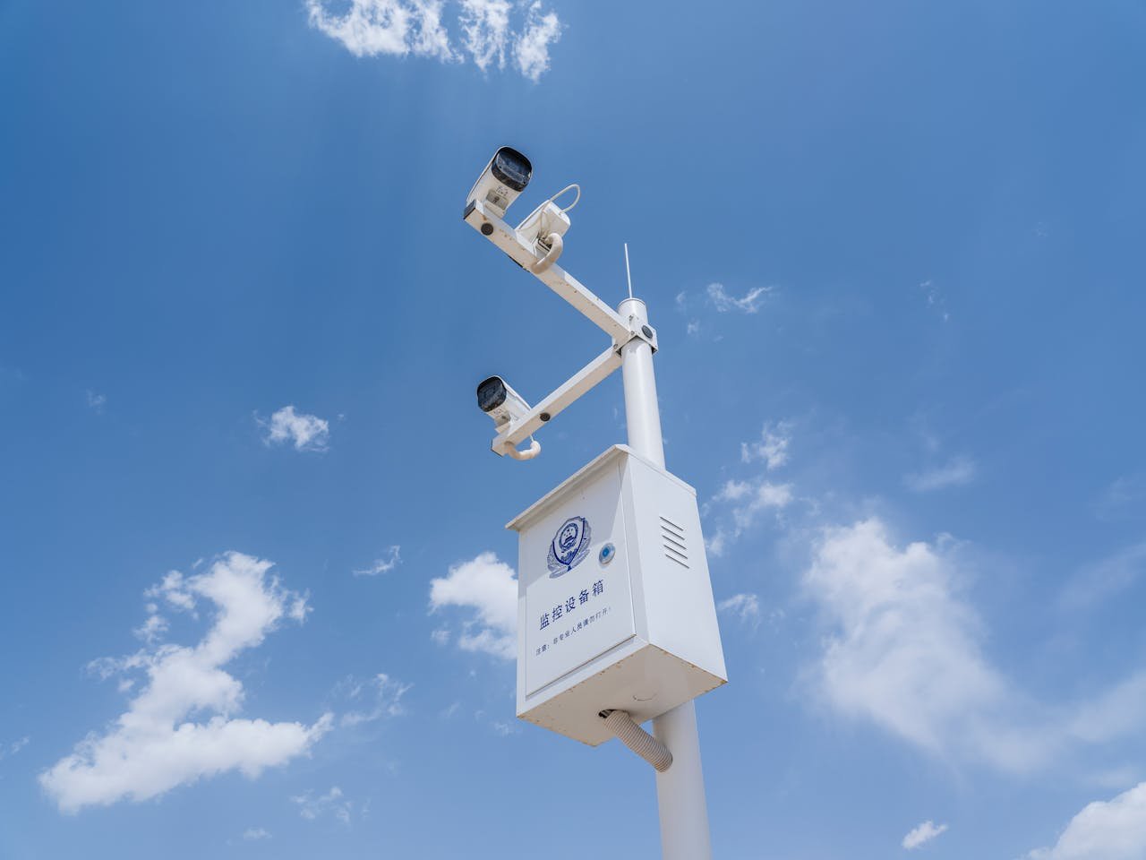 White security cameras mounted on a pole against a bright blue sky with fluffy clouds.